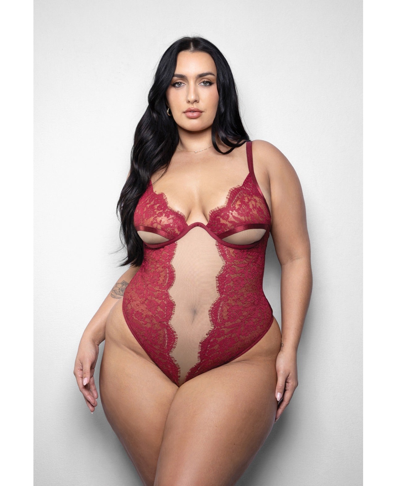 A plus-size model wearing a red lace and mesh cut-out bodysuit, standing against a light gray studio background.