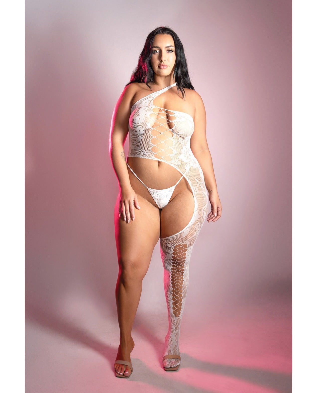 A woman modeling a white one-shoulder lace bodysuit with a fishnet thigh-high stocking and matching white underwear, standing against a pink studio backdrop.