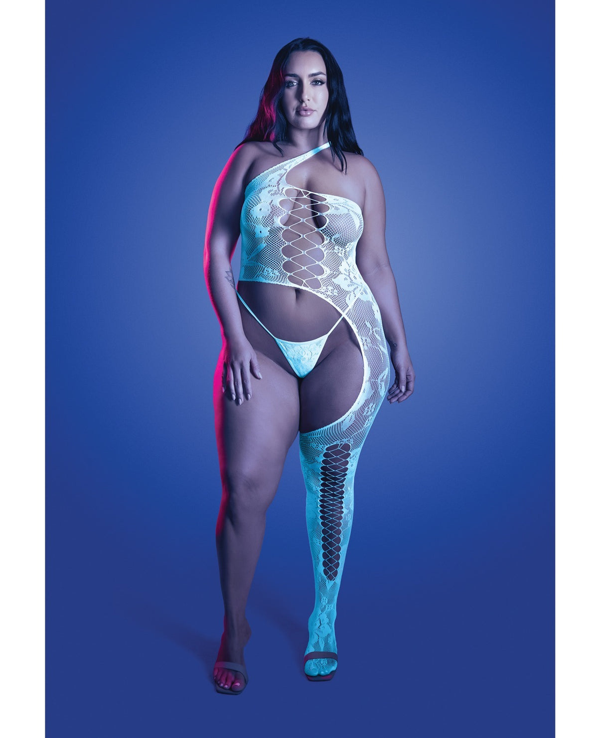 A woman modeling a sheer, one-shoulder bodysuit and matching thong with integrated thigh-high stockings against a blue studio background.