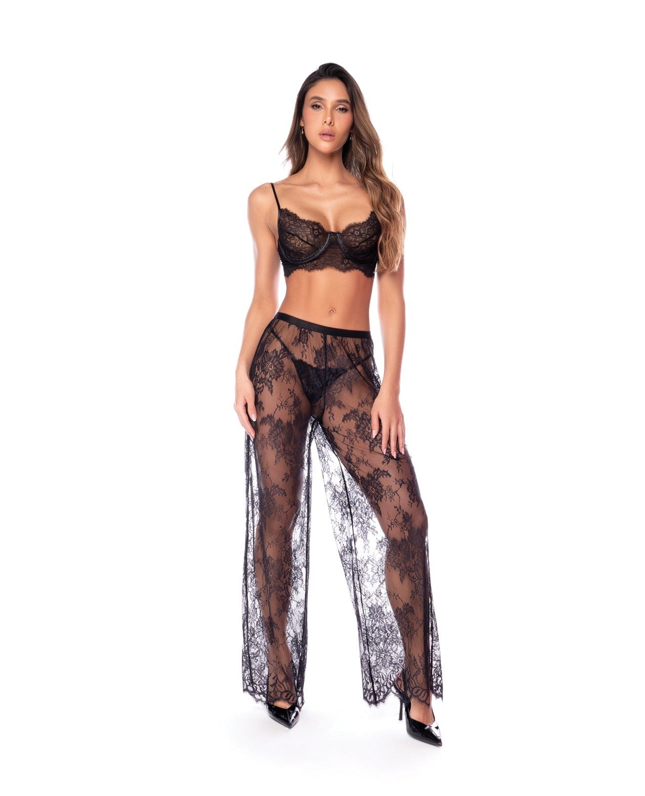 A woman modeling a black sheer lace lingerie set consisting of a bra, matching sheer pants, and black pointed-toe heels against a plain white background.