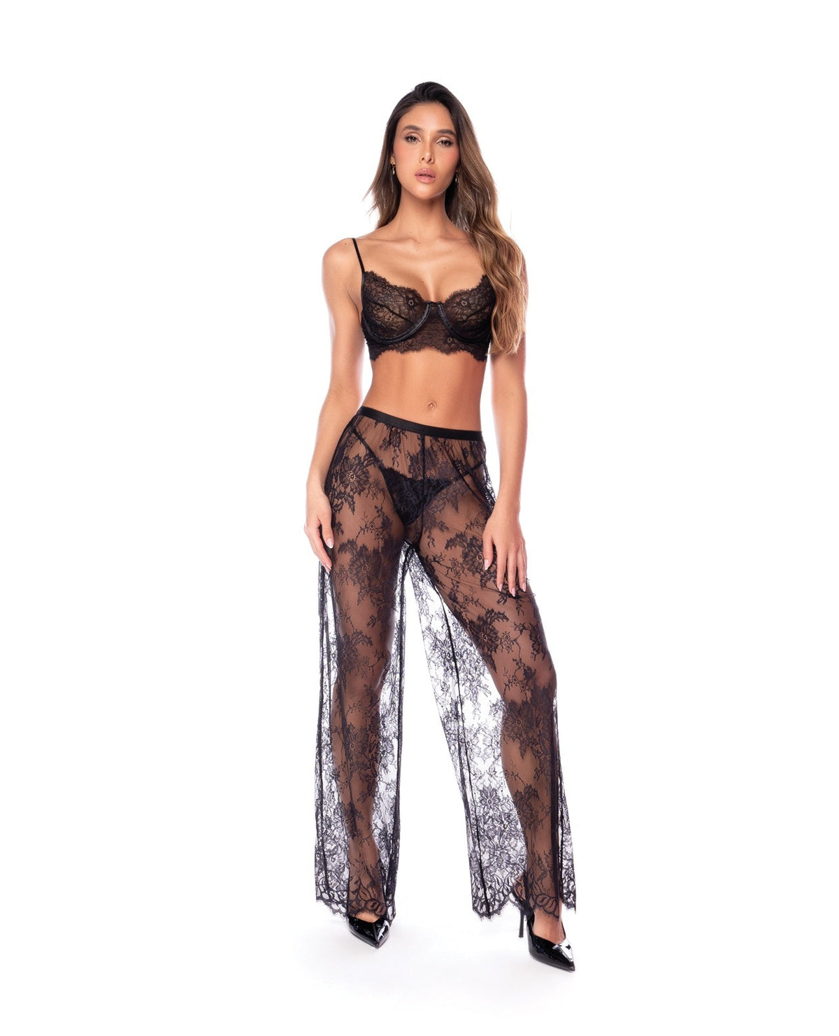 A woman modeling a black sheer lace lingerie set consisting of a bra, matching sheer pants, and black pointed-toe heels against a plain white background.