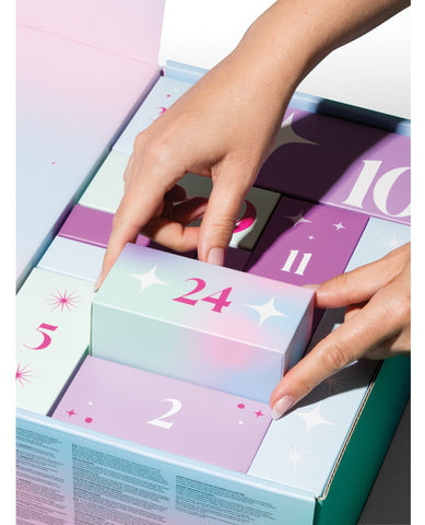 A close-up of hands opening an advent calendar box, lifting the front flap marked 24. The box contains numbered pastel compartments in mint, lavender, and purple tones with star and number illustrations.